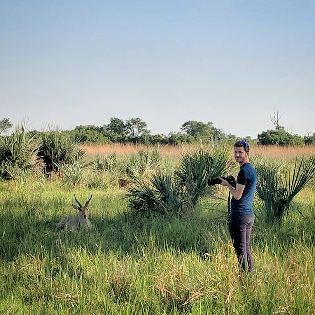 Maxx with a camera in long grass, photographing an antelope lying a short distance away